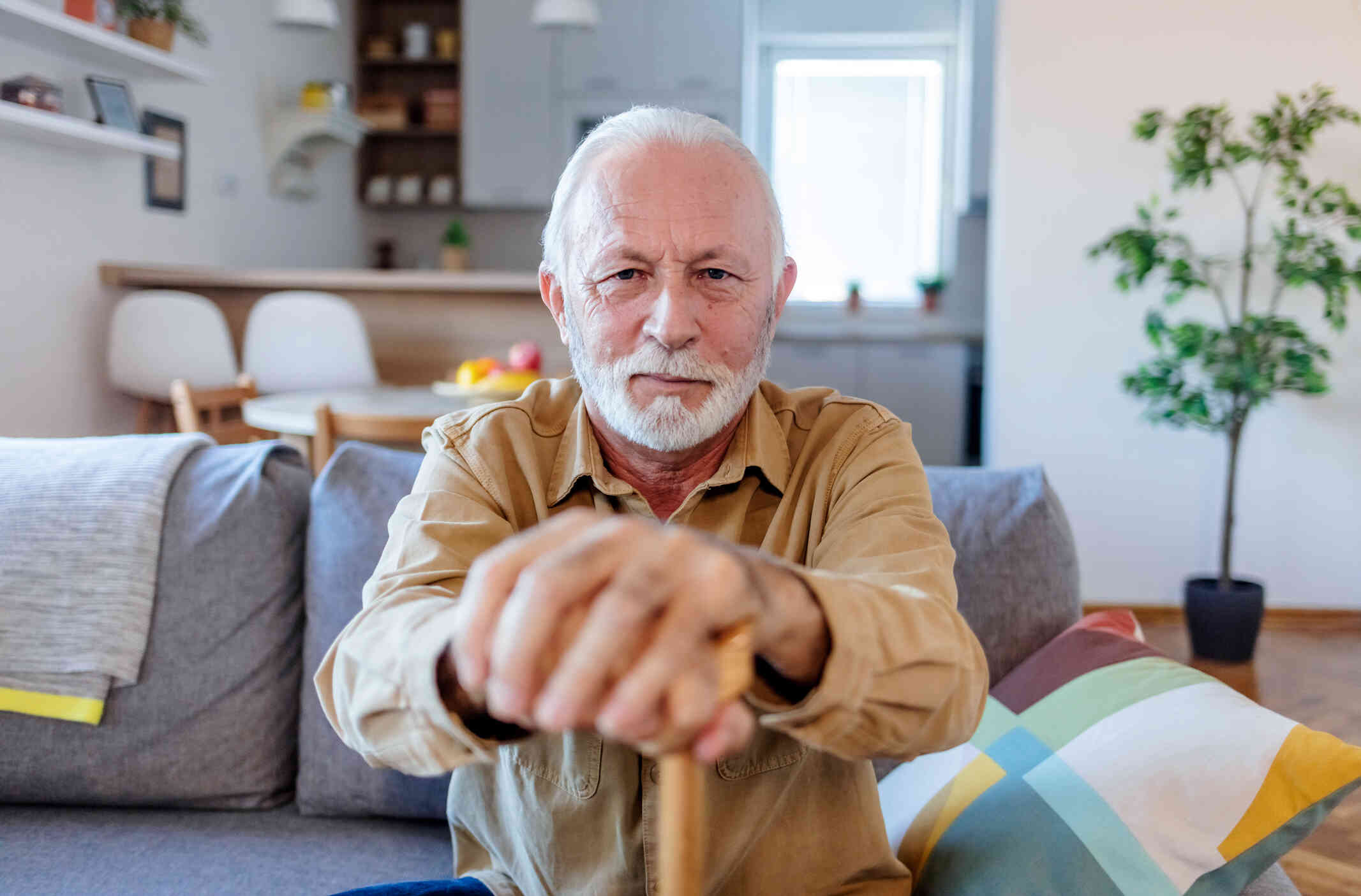 An elderly man in a button down shirt sits on the couch with his cane in his hands while looking at the camera with a soft smile.